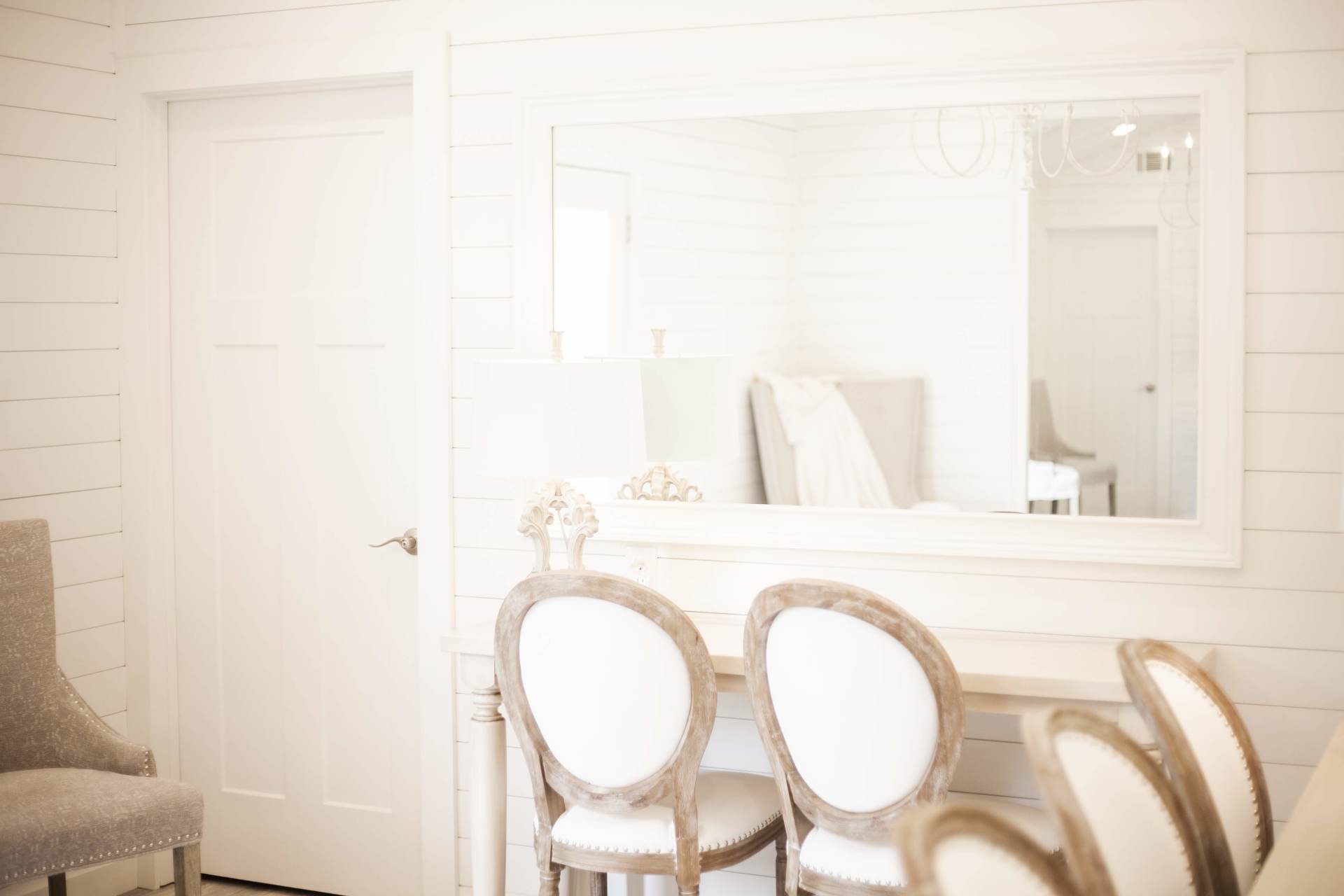 Bright white vanity area with large mirror, door, and three ornate chairs in a sunlit room