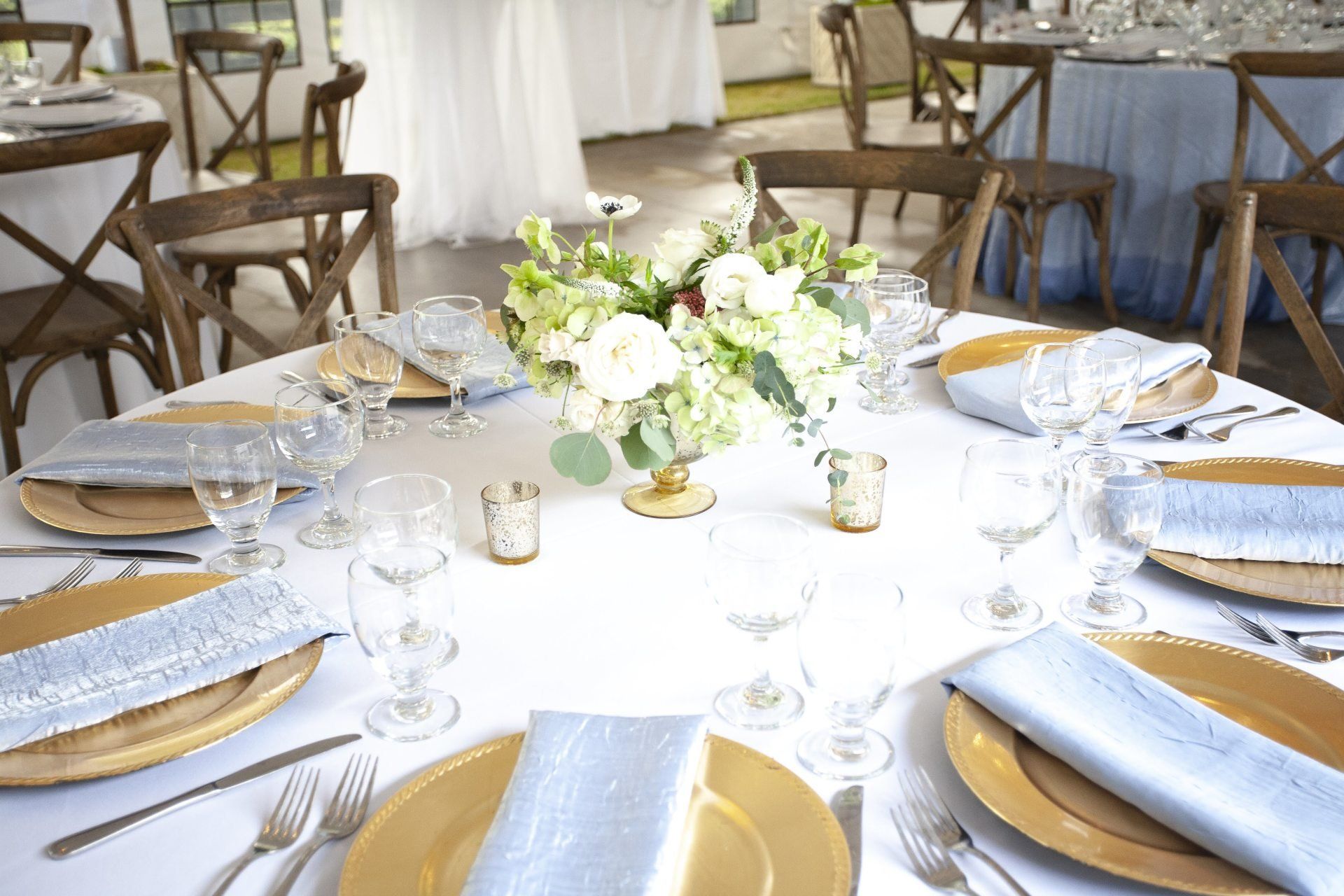 Round banquet table set with gold chargers, glassware, and a white flower centerpiece in a bright reception hall
