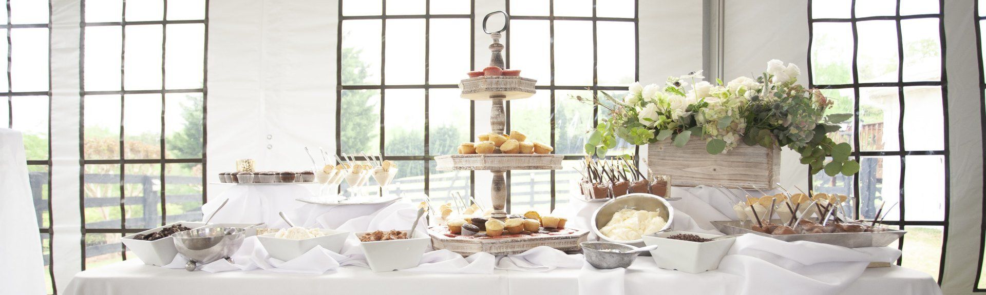 Elegant dessert table by large windows with white flowers, cakes, and table settings