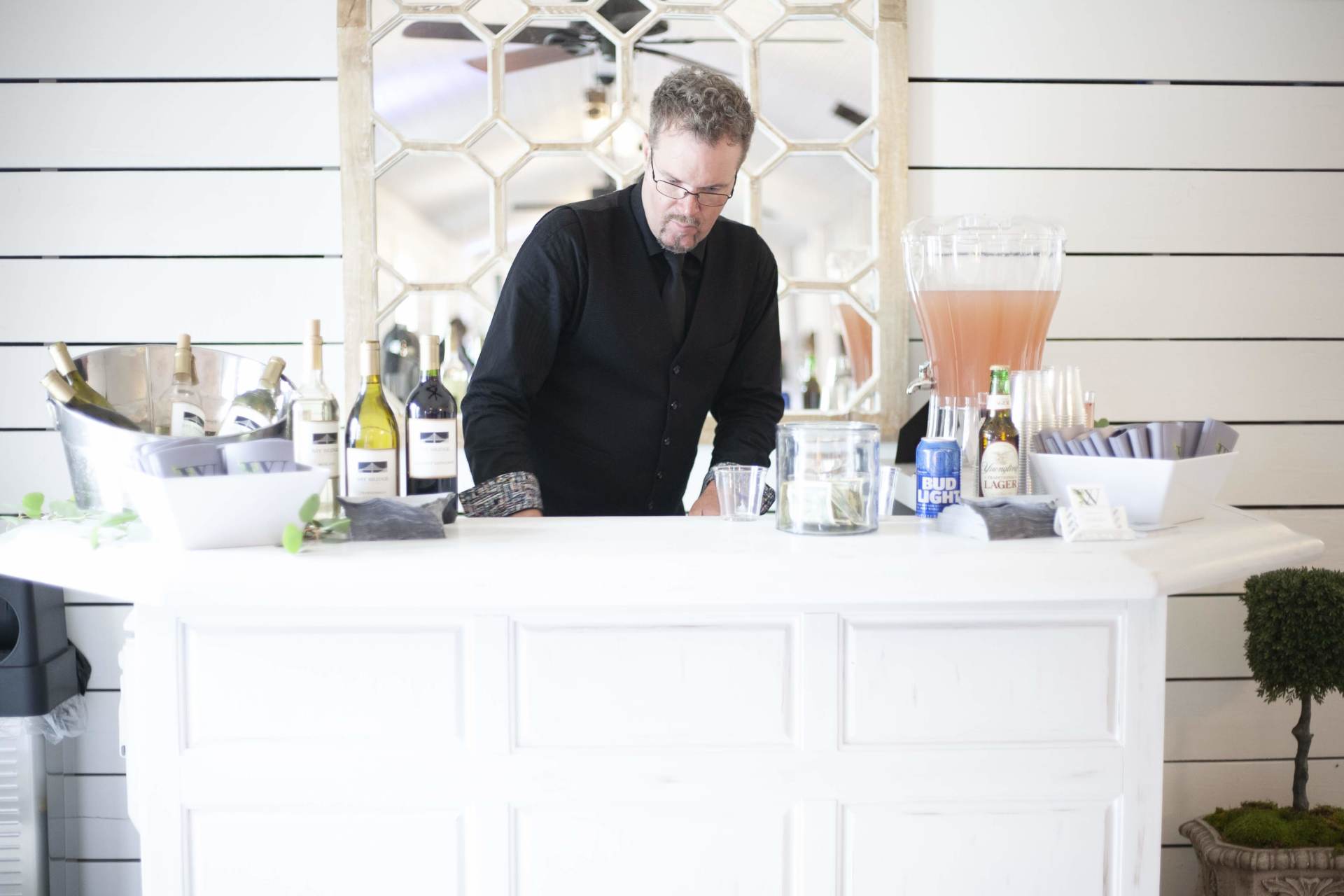 Bartender in a black shirt mixing drinks at a white bar with bottles and cocktail ingredients