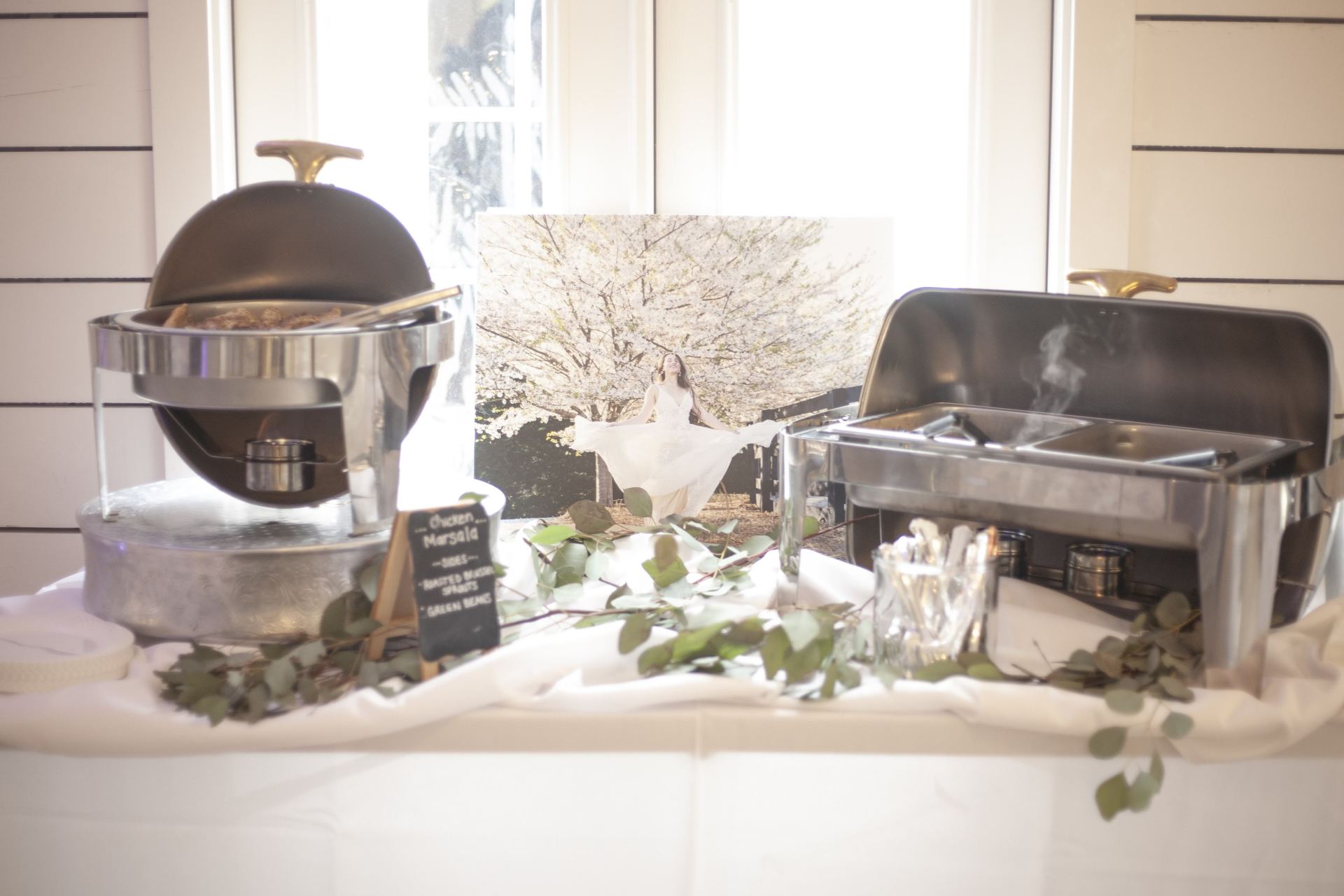 Buffet table with chafing dishes, serving utensils, and greenery on a white tablecloth