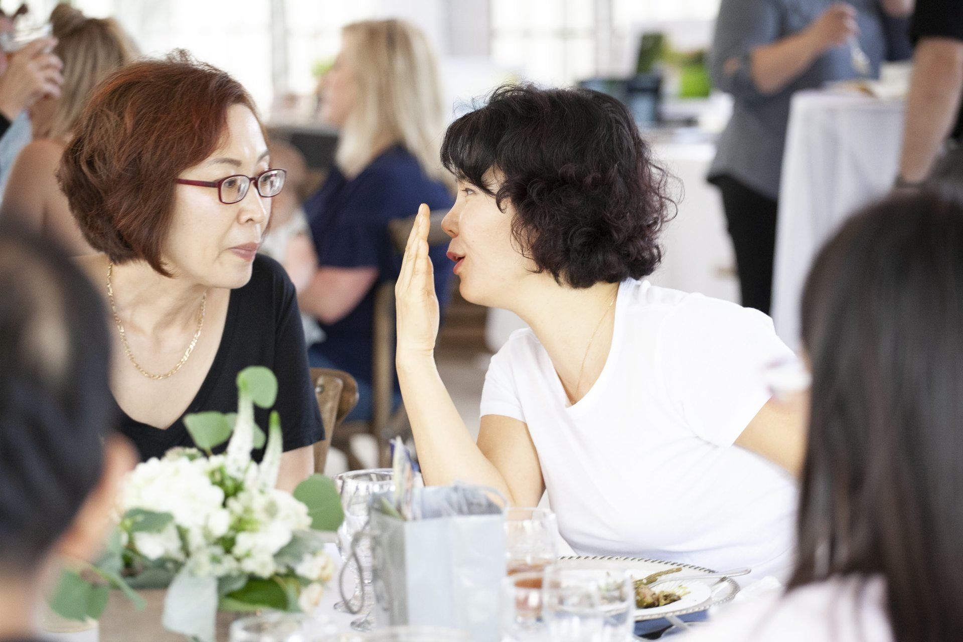 Two women talking at a table during a social event, with flowers and drinks in the foreground.