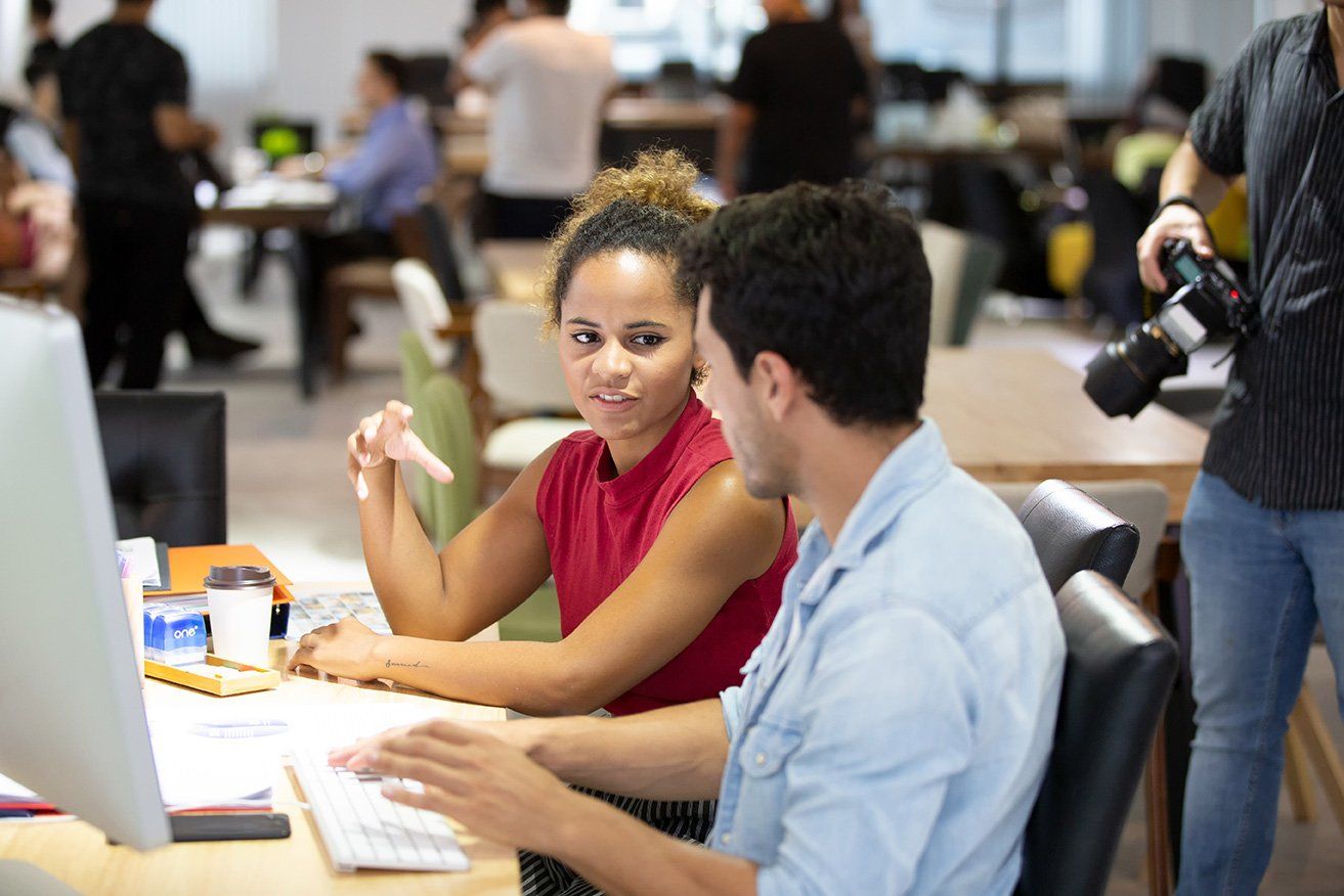 A man and a woman are sitting at a desk looking at a computer screen.