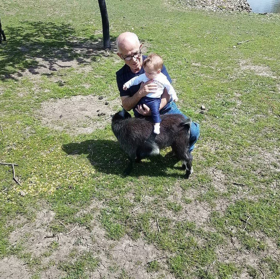 A man is holding a baby while sitting on a sheep.