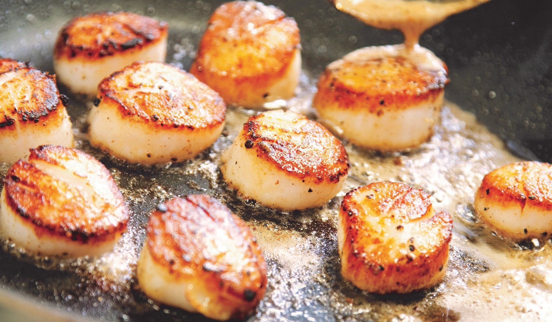 Scallops are being cooked in a pan with sauce being poured on them.