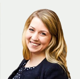 A smiling person with long blonde hair, wearing a dark blue jacket over a polka-dot shirt, against a plain background.
