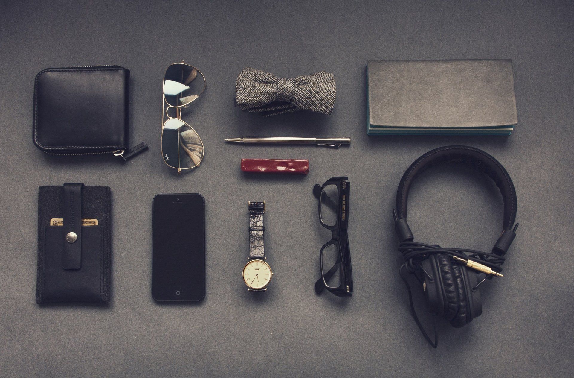 Flat lay of men's personal accessories, including a wallet, sunglasses, watch, headphones, and small items on a dark surface.