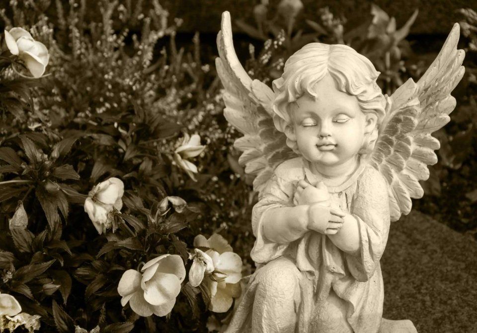 Statua di un angelo in pietra inginocchiato con una colomba, circondato da fiori in un giardino.