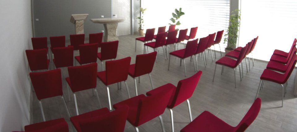 Una piccola sala moderna con sedie rosse, ideale per riunioni o cerimonie. Un podio e un tavolo si trovano nella parte anteriore.