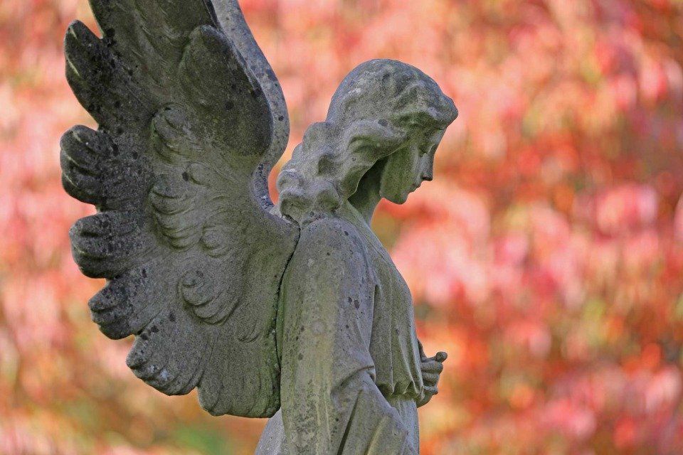 Statua di un angelo in pietra con il viso abbassato e le ali spiegate, su uno sfondo autunnale sfocato di colore arancione e rosso.