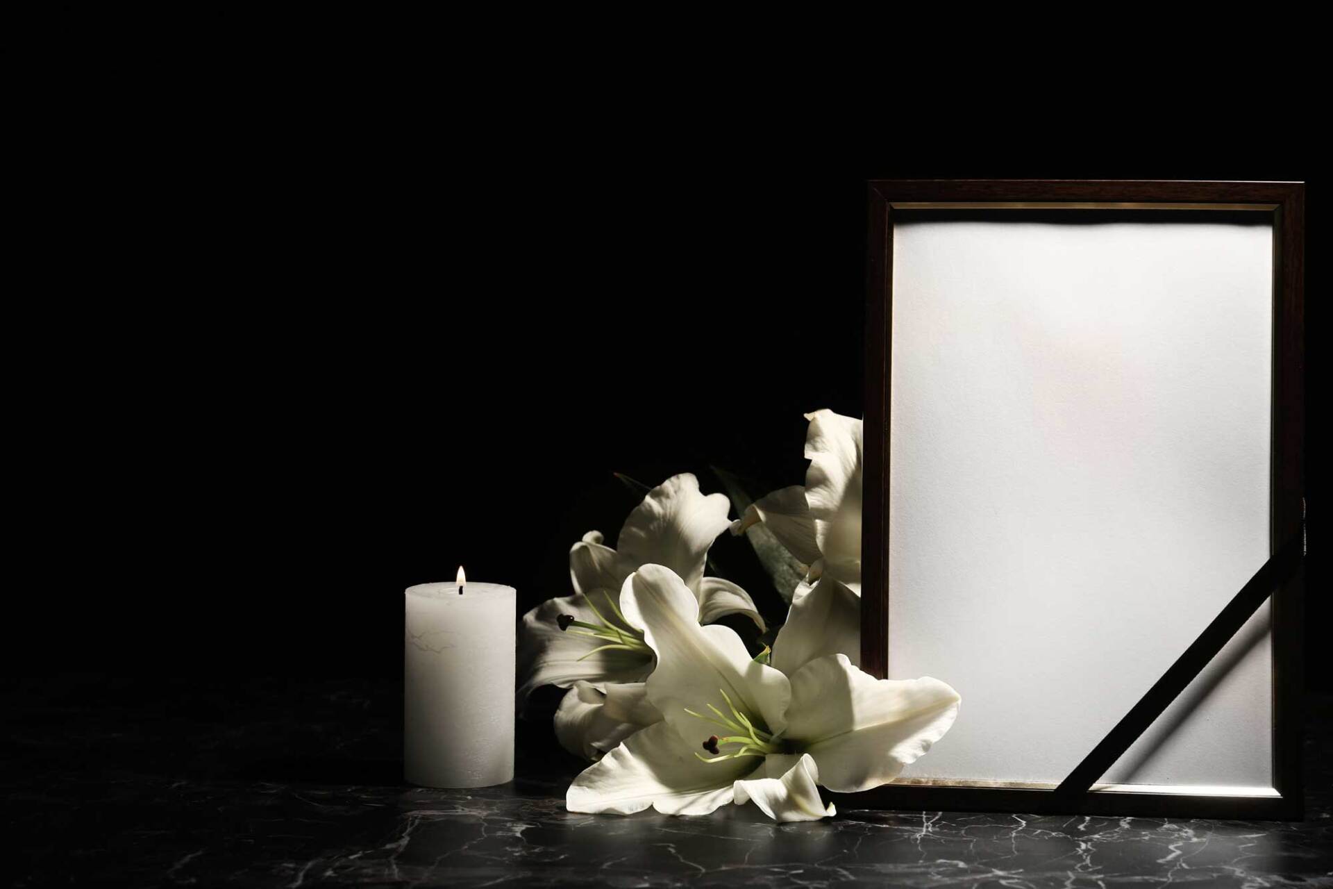 Candela, gigli bianchi e cornice vuota con nastro nero, su sfondo nero.