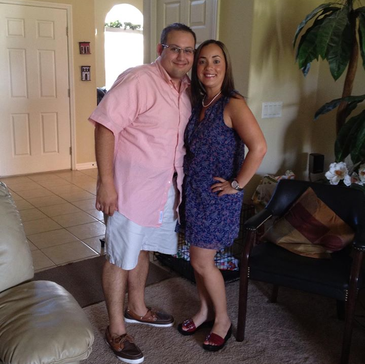 A man and woman smiling in an indoor setting, wearing casual clothes and standing side-by-side.