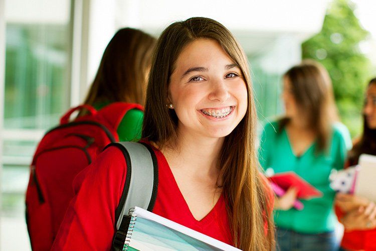 Teenage girl with braces smiles at the camera, holding a notebook and wearing a red shirt, classmates blurred in the background.