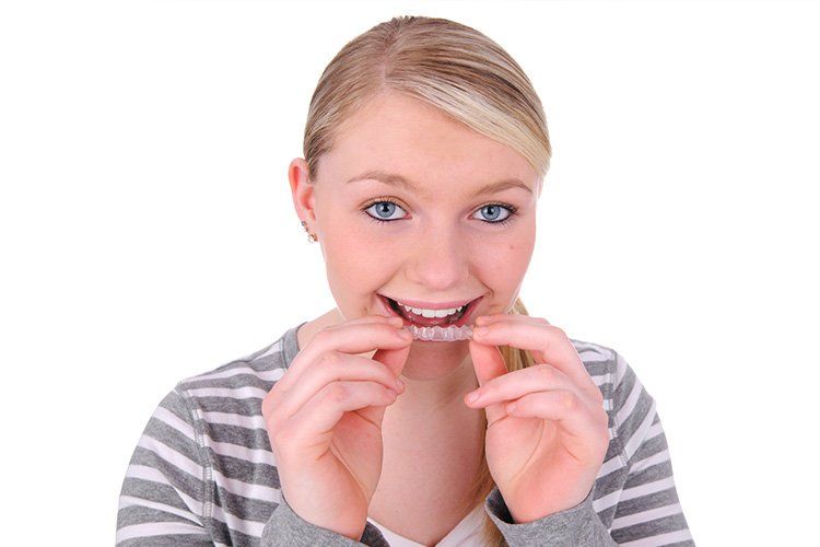 Woman with blonde hair smiling, holding clear Invisalign aligner in her hands.
