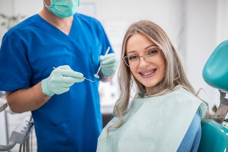 Woman with braces smiles at the camera in a dentist's chair; dentist in blue scrubs stands behind her.