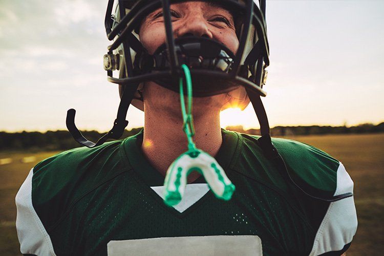 Football player in a green jersey with helmet, pulling down mouthguard against a sunset backdrop.