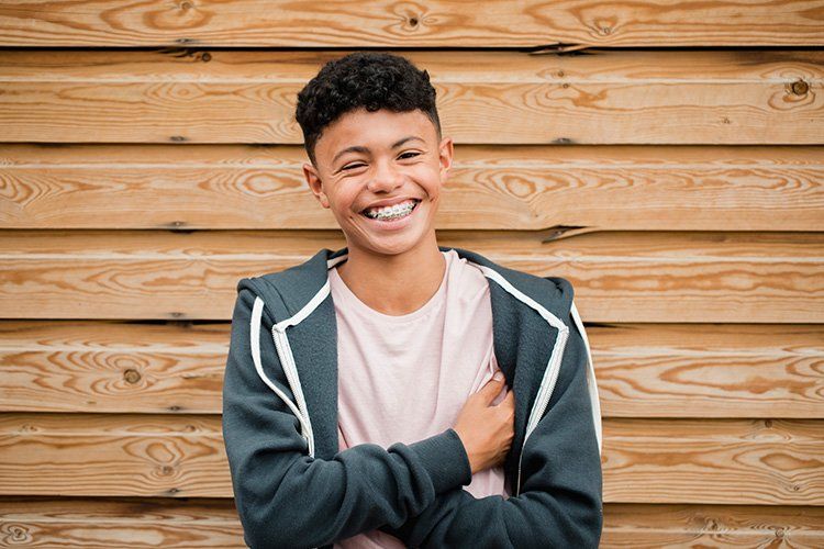 Teen boy with curly hair smiles broadly, arms crossed in front of a wood-paneled wall.