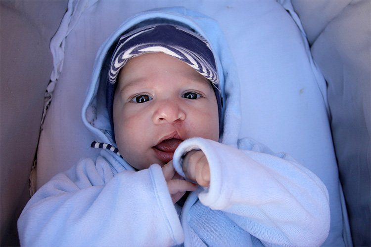 Baby in blue hooded onesie and hat in stroller, looking at the camera with mouth slightly open.