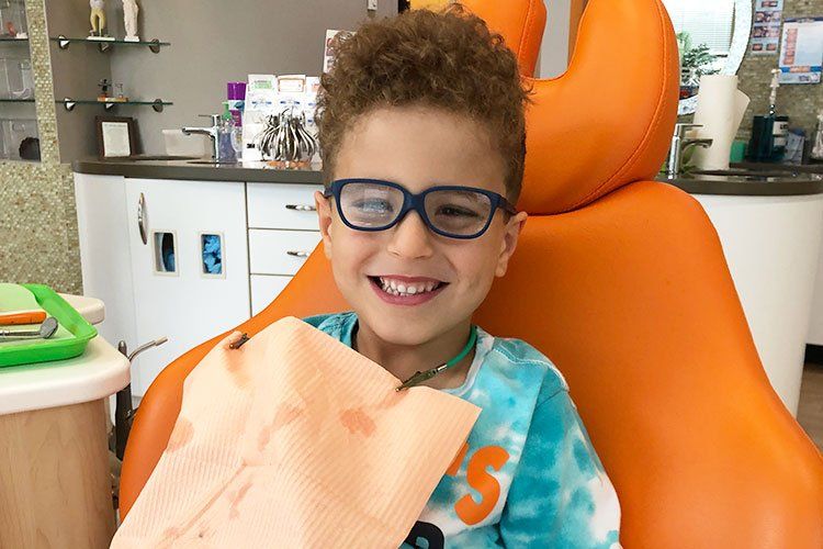 Smiling child in glasses sitting in an orange dental chair, with a bib.
