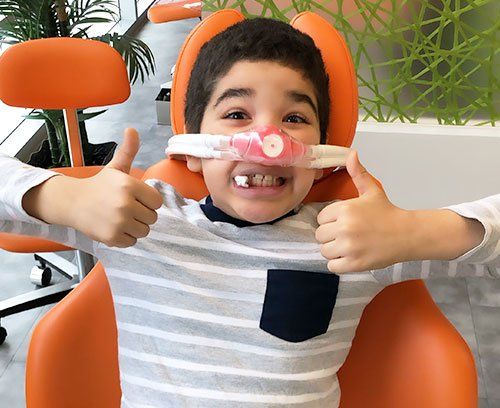 Boy in dental chair with nasal mask giving thumbs-up. Orange chair, striped shirt, happy expression.