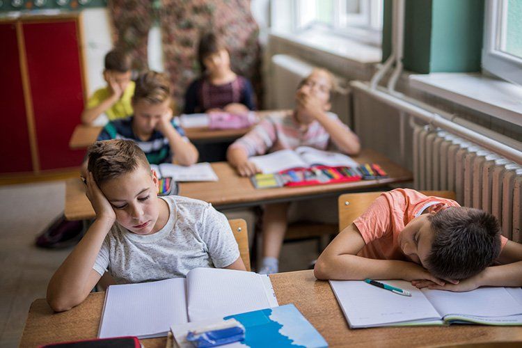 Tired students in a classroom, resting heads on desks, books open.