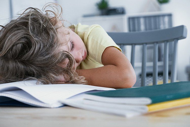 Tired child resting head on a desk over open books, in a light-filled room.