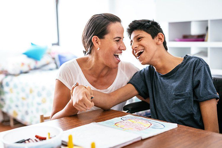 Woman and boy laughing together, drawing at a table. Bright room, crayons, and bed in background.