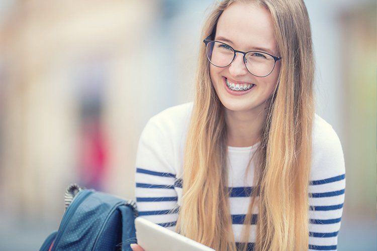 Blonde woman with glasses and braces smiles, holding a tablet. She wears a striped shirt, with a backpack nearby.