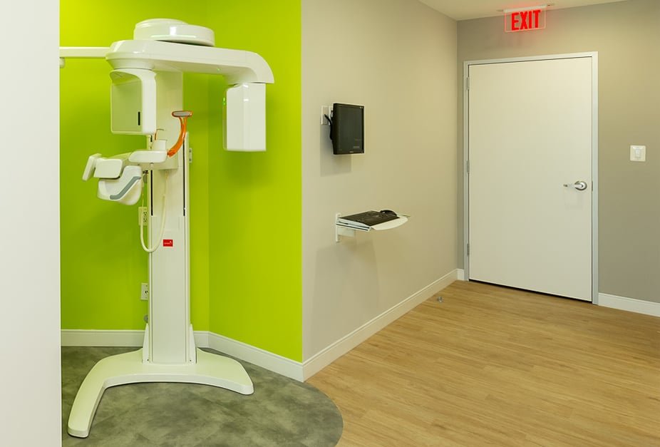 Dental office with panoramic X-ray machine. Green and gray walls, white door, and wood-look floor.