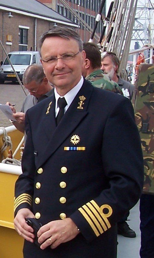 Man in marine-uniform, staand op een schip, glimlachend. Gouden insignes, gouden strepen op de mouwen.