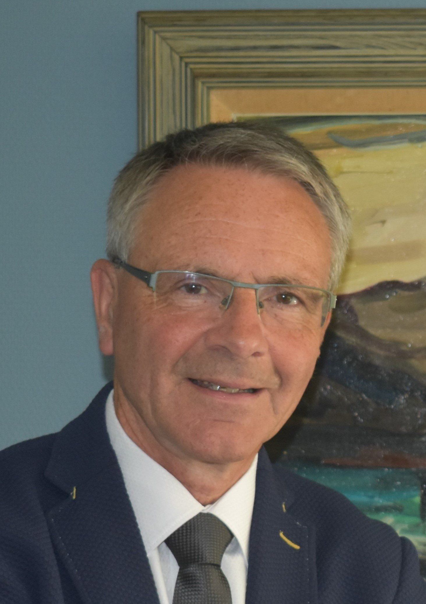 Een man met bril en pak glimlacht, staand tegen een muur met ingelijste kunstwerken.