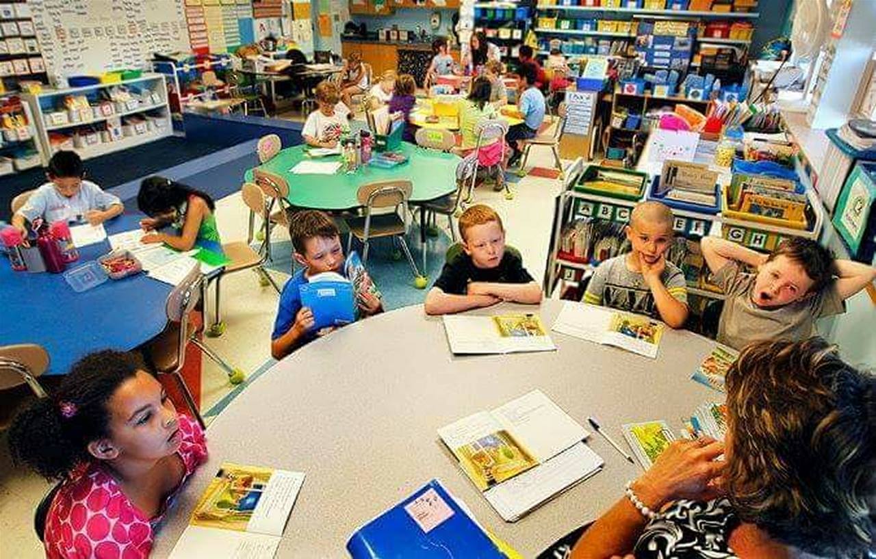 Children in a classroom seated at tables, some reading, with a teacher. Bright setting with bookshelves.