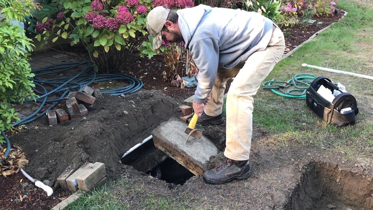 A person in work clothes uses a small hammer to lift a concrete cover off an underground utility box in a garden.