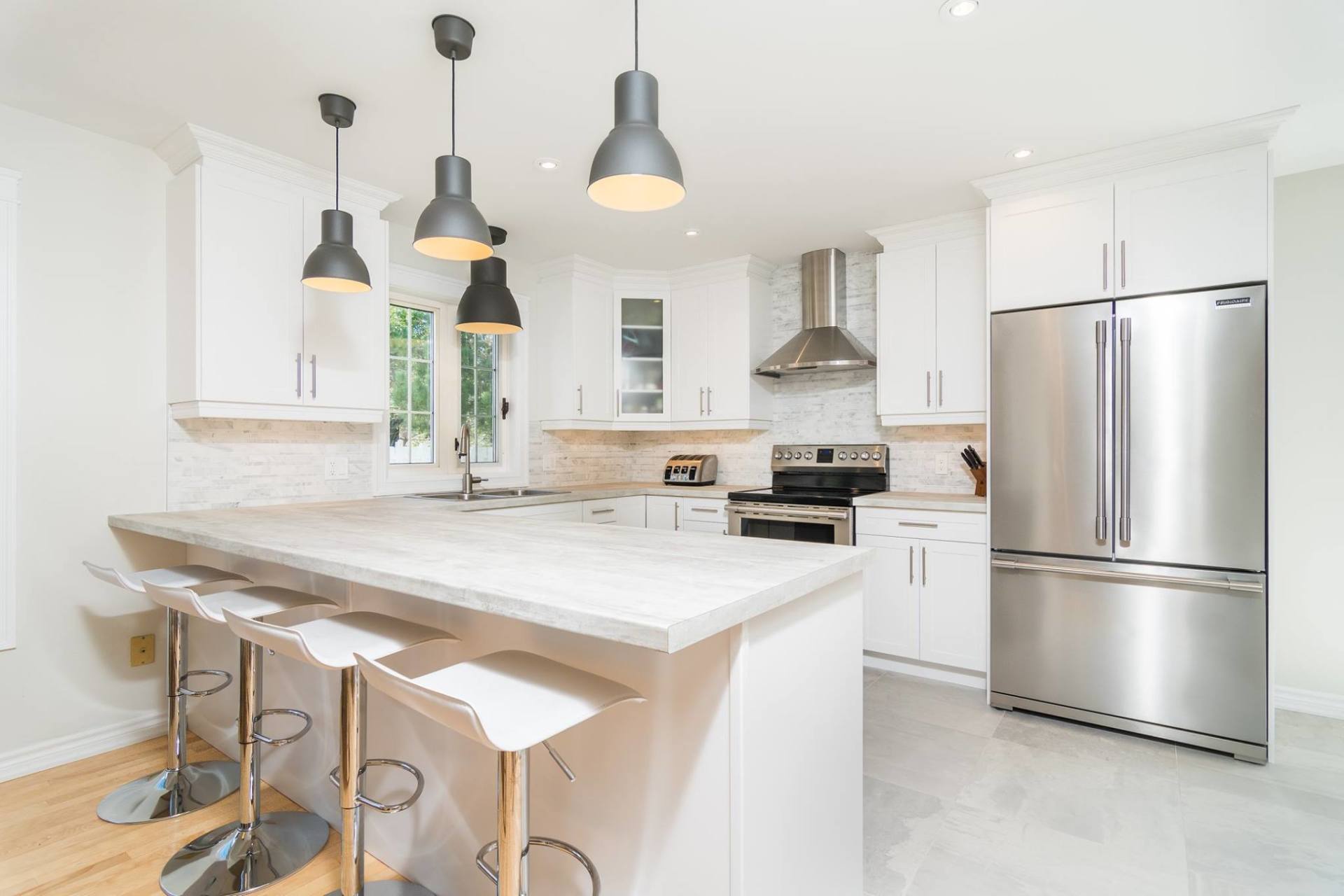 Modern white kitchen with a granite island, stainless steel appliances, and three bar stools under hanging lights.