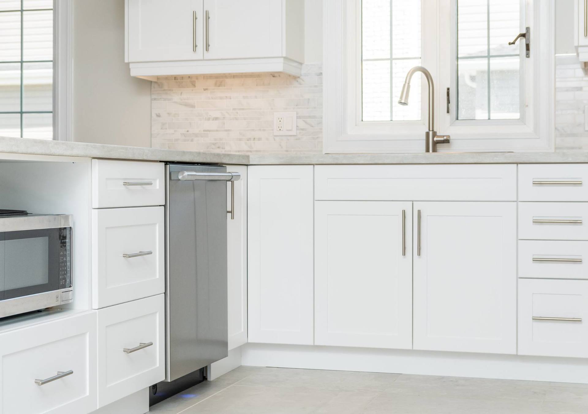A modern kitchen with white cabinets, a stainless steel dishwasher and microwave, and a chrome faucet under a window.