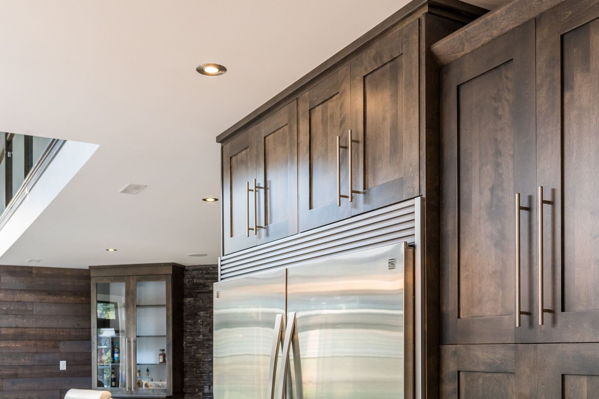 Modern dark wood kitchen cabinetry surrounding a stainless steel refrigerator in a well-lit home.