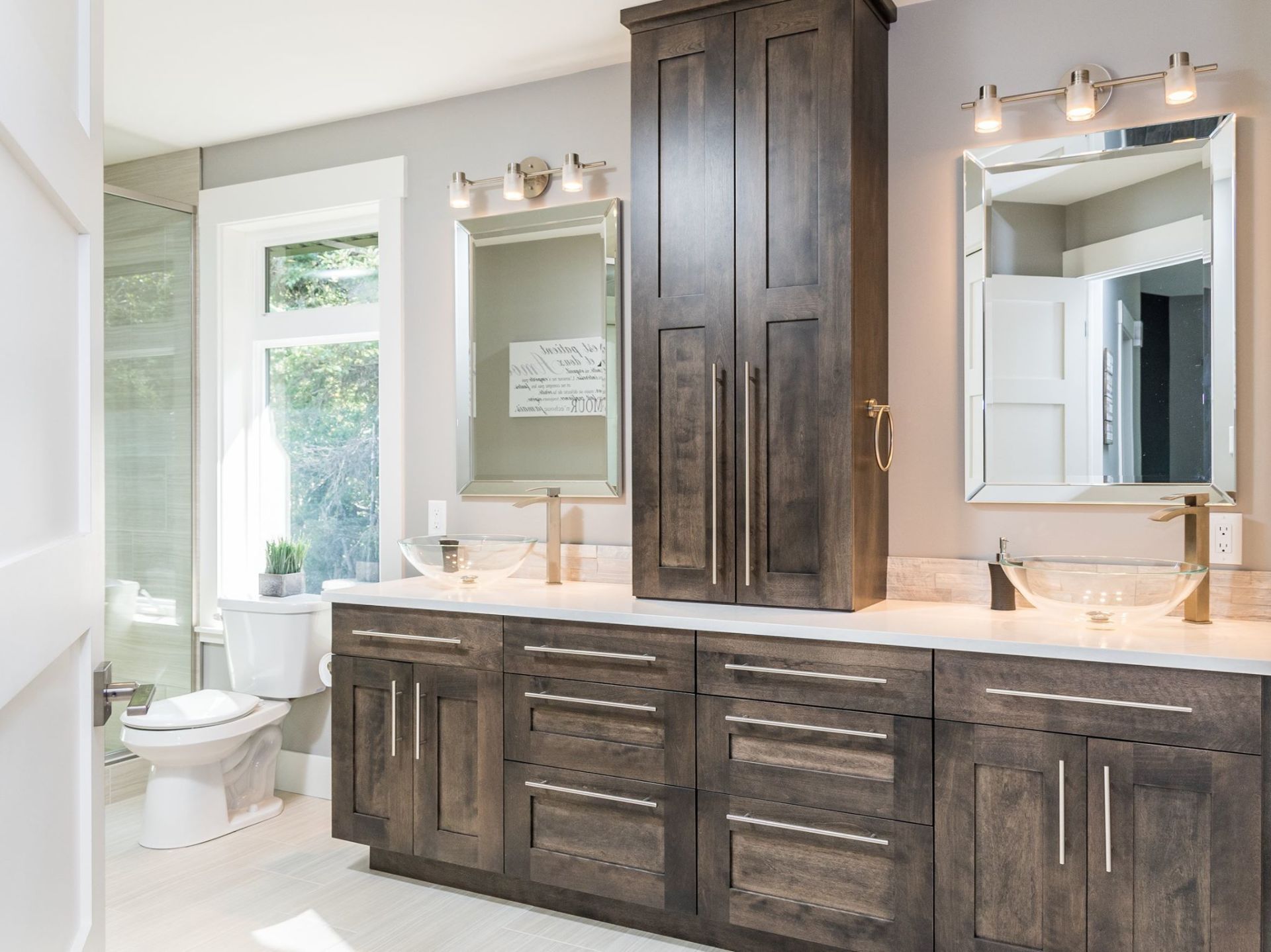 A modern bathroom featuring a double vanity with dark wood cabinets, a central tower, two mirrors, and gold fixtures.