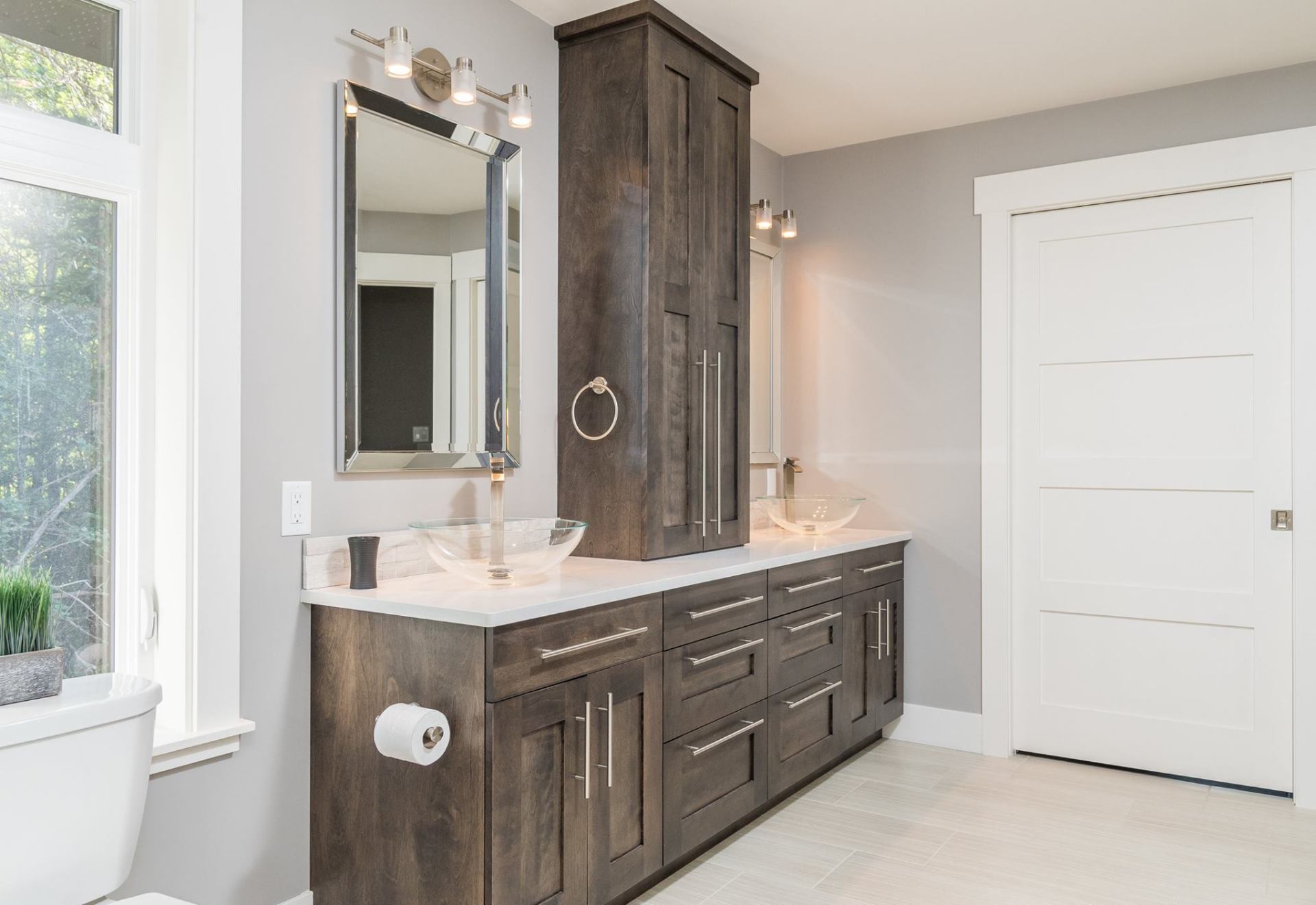 A modern bathroom featuring a dark wood vanity with a white countertop, two vessel sinks, a tall cabinet, and gray walls.