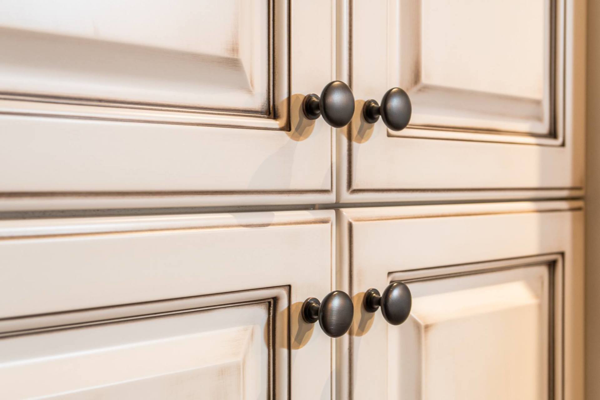 Close-up of off-white kitchen cabinet doors with a distressed glaze finish and dark round metal knobs.