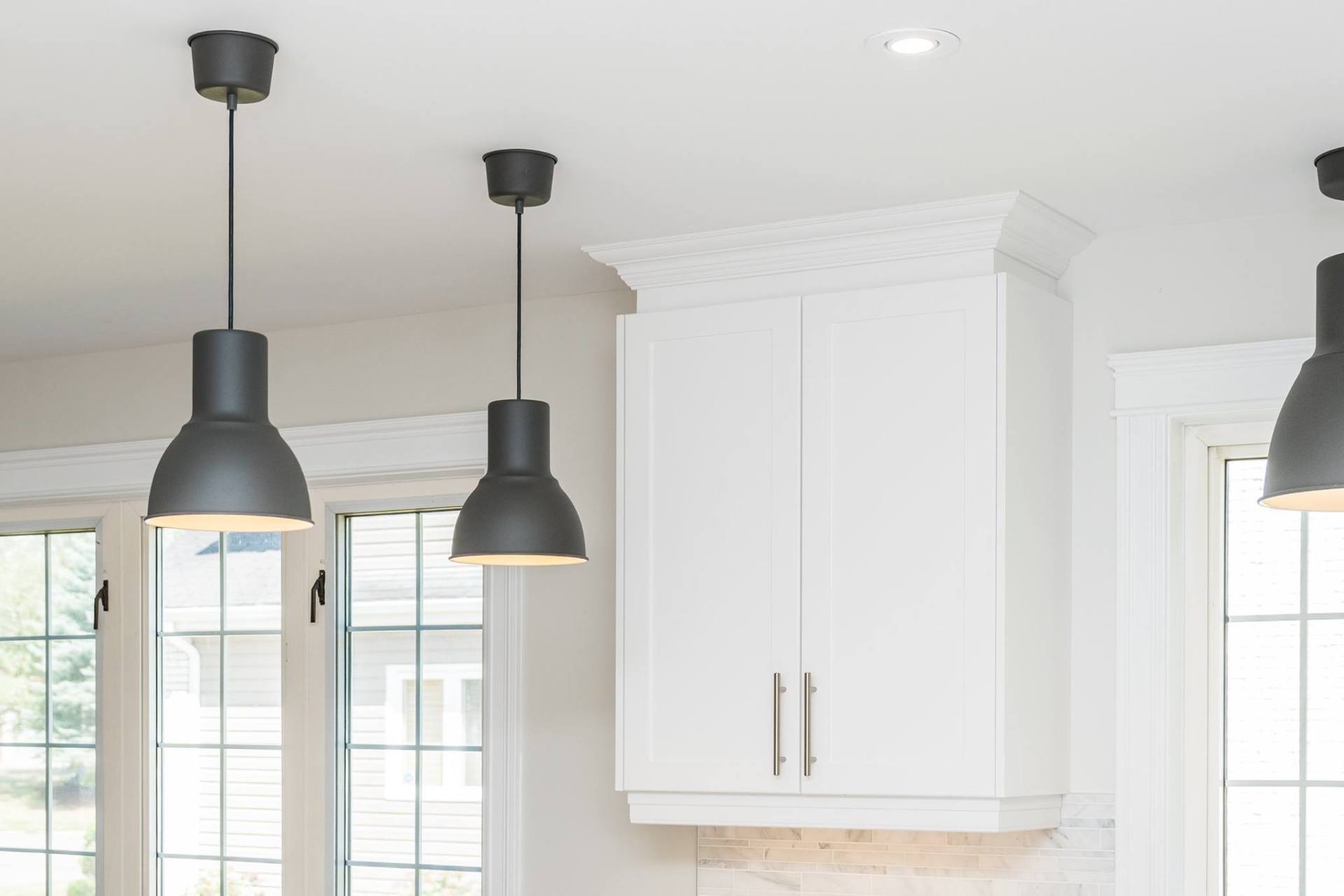 Three matte black pendant lights hang from a white ceiling beside a white kitchen cabinet with crown molding.