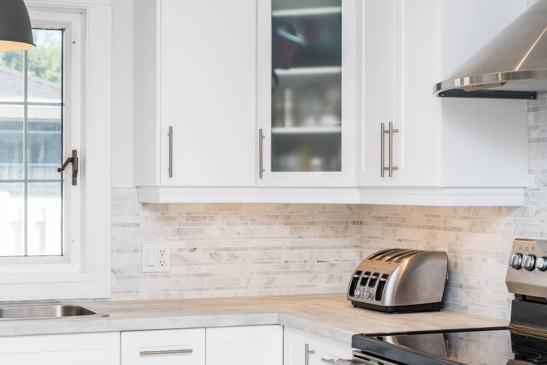 A modern kitchen featuring white cabinets, a light-colored brick backsplash, stainless steel appliances, and a window.