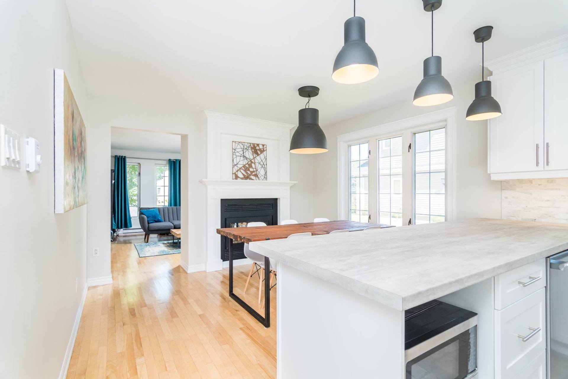 A bright, modern kitchen and dining area with white cabinets, a light stone island, wood floors, and hanging black lights.