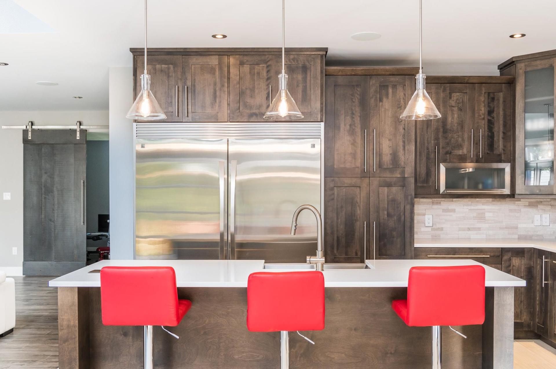 Modern kitchen with dark wood cabinets, stainless steel refrigerator, white countertops, and three red bar stools.