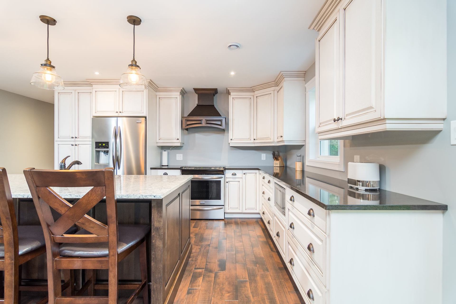 A kitchen with cream cabinets, stainless steel appliances, a dark wooden island with two chairs, and hardwood flooring.