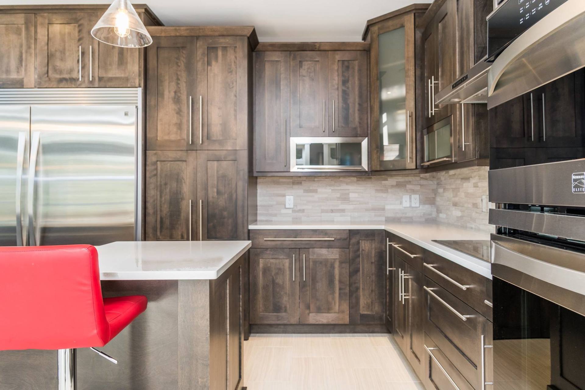 Modern kitchen with dark wood cabinets, stainless steel appliances, white countertops, and a single red bar stool.