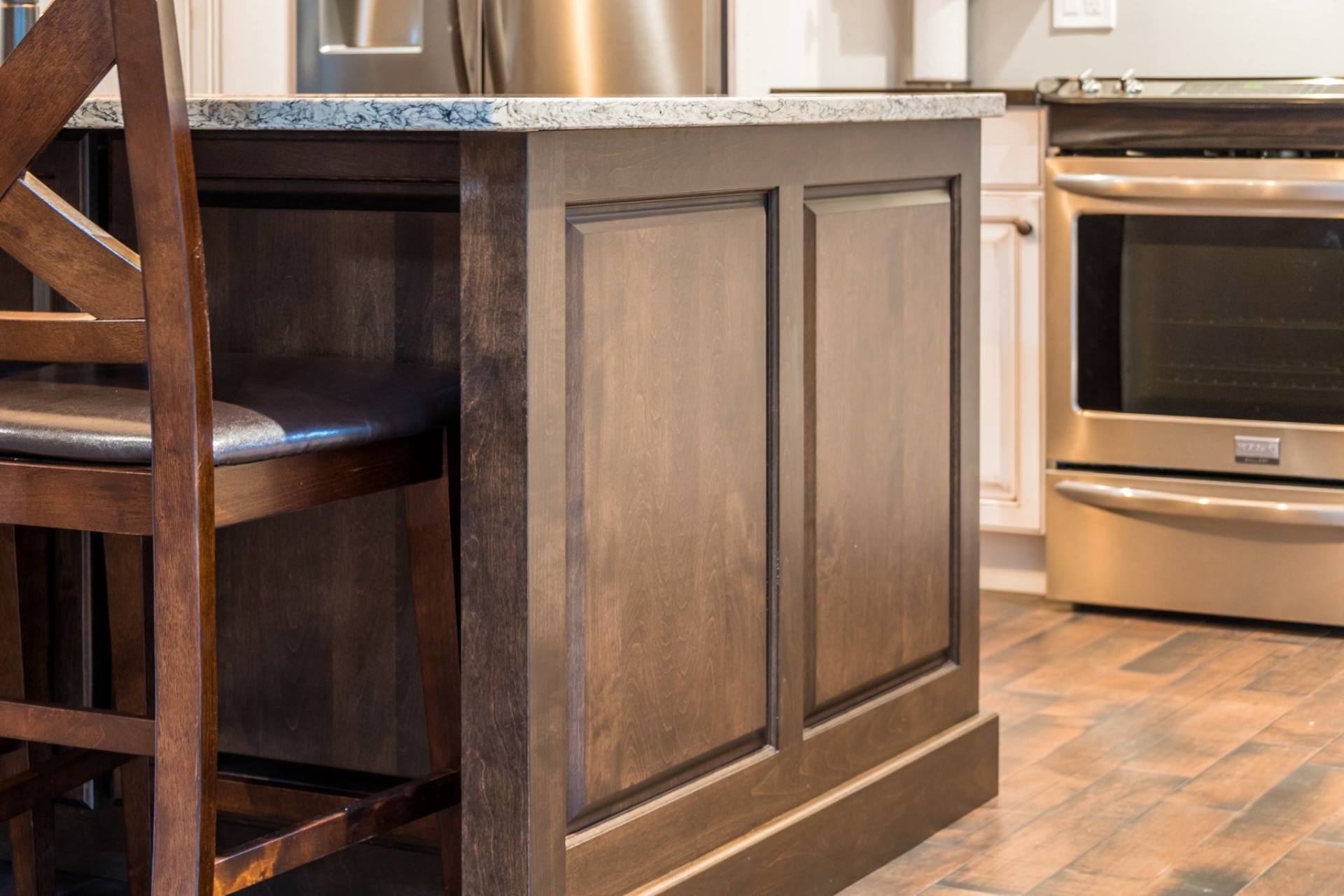 A wooden kitchen island with a granite countertop, featuring recessed paneling and a nearby stool and stainless oven.