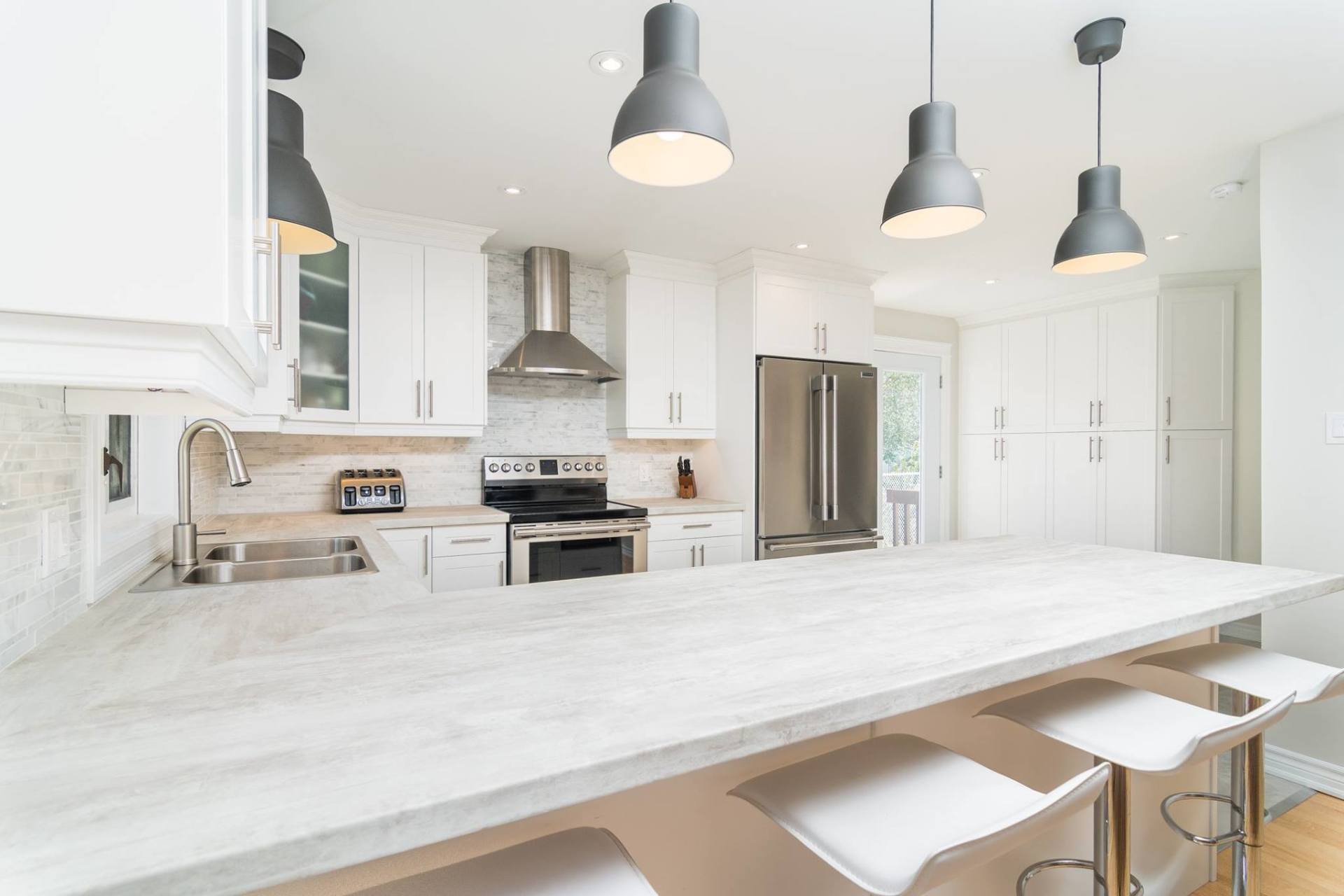 A modern, brightly lit kitchen with white cabinetry, a light-toned marble island, and three gray pendant lights.