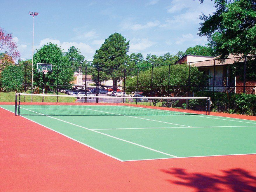 Tennis court with green playing surface, red surround, and net. Buildings and trees in the background.