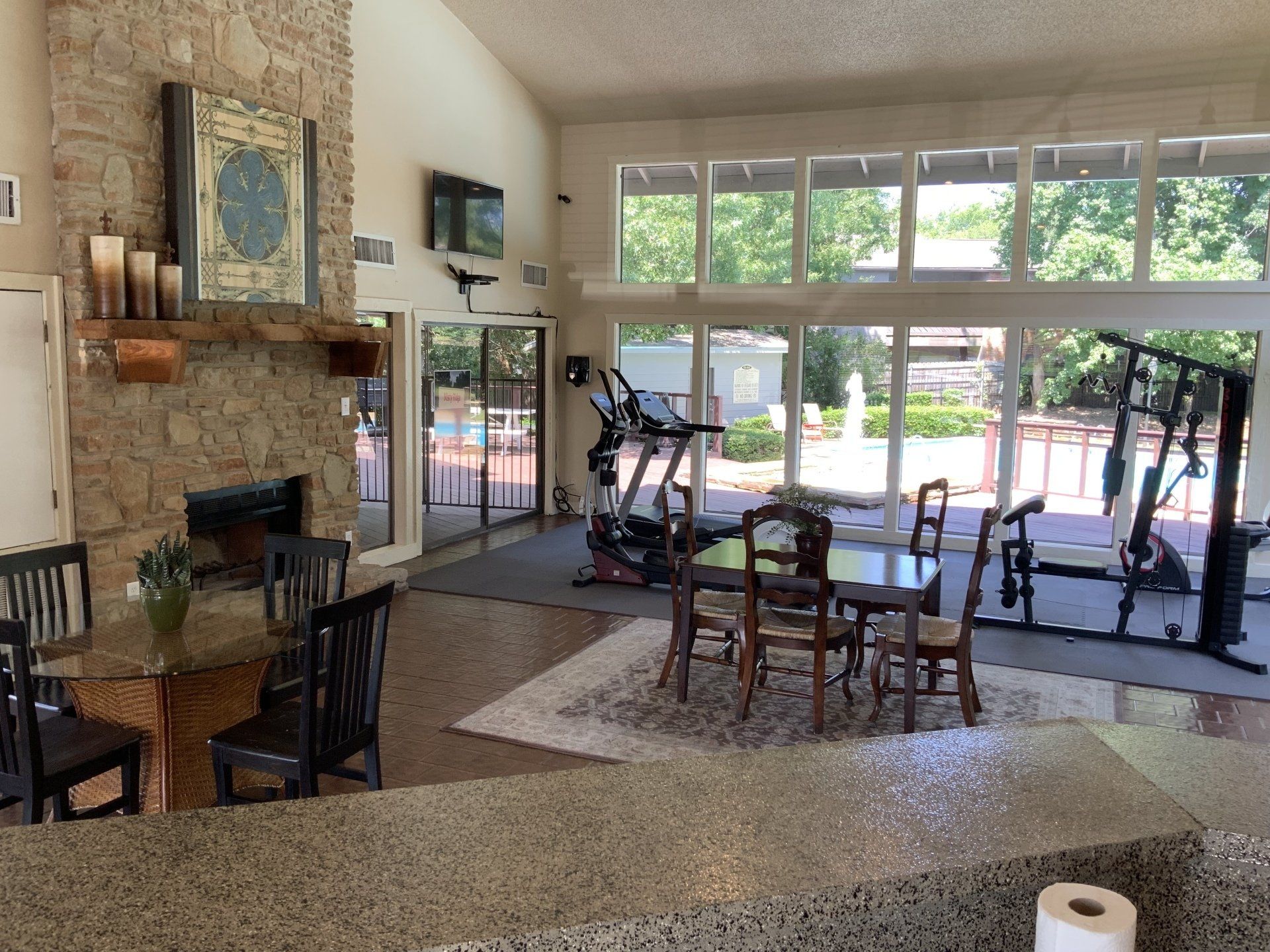 Interior view of a clubhouse with exercise equipment, a fireplace, and a dining area. Large windows look out to a patio.