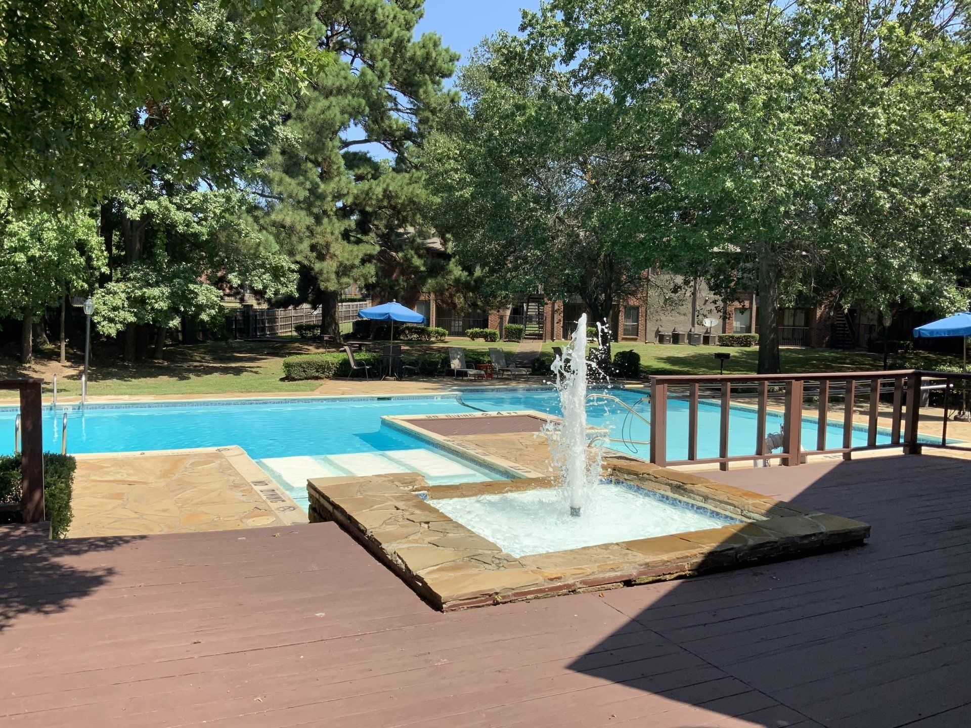 Pool area with fountain, deck, and trees on a sunny day.